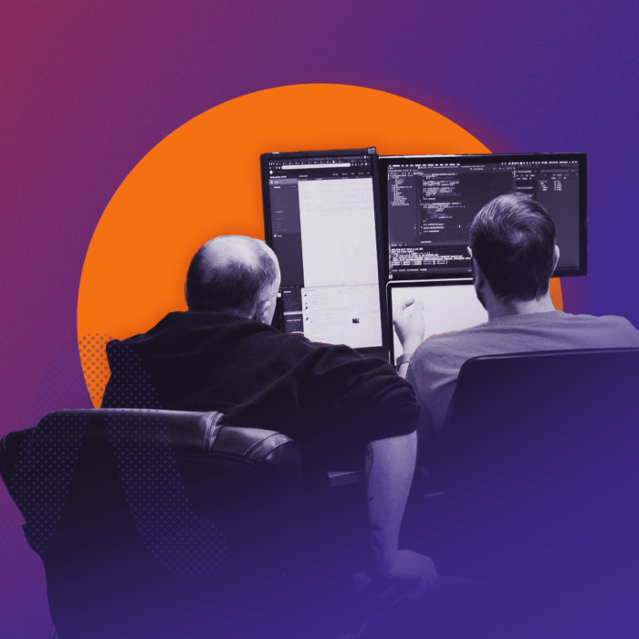 Two men sit facing multiple desktop monitors