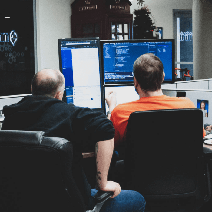 Two people sitting at a desk and facing a vertical computer monitor, a horizontal computer monitor, and a laptop, working together to solve a problem.
