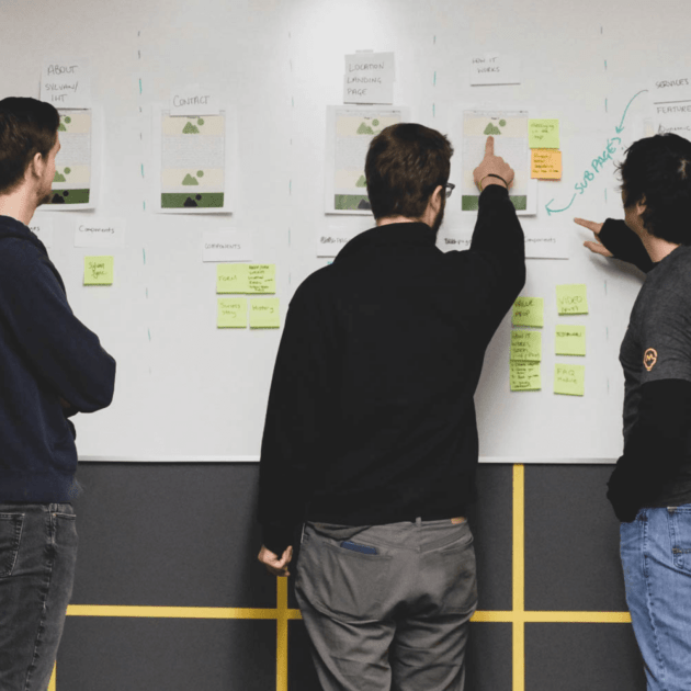 Three people looking at papers and notes on a white board, two of the people are pointing at a specific page on the right