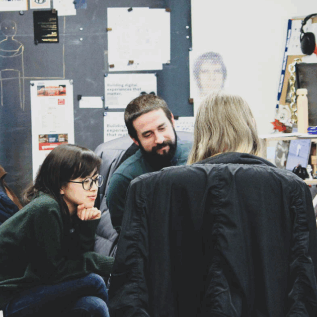 Three people sitting down and facing each other. Two people face the camera looking at the person facing away from the camera.
