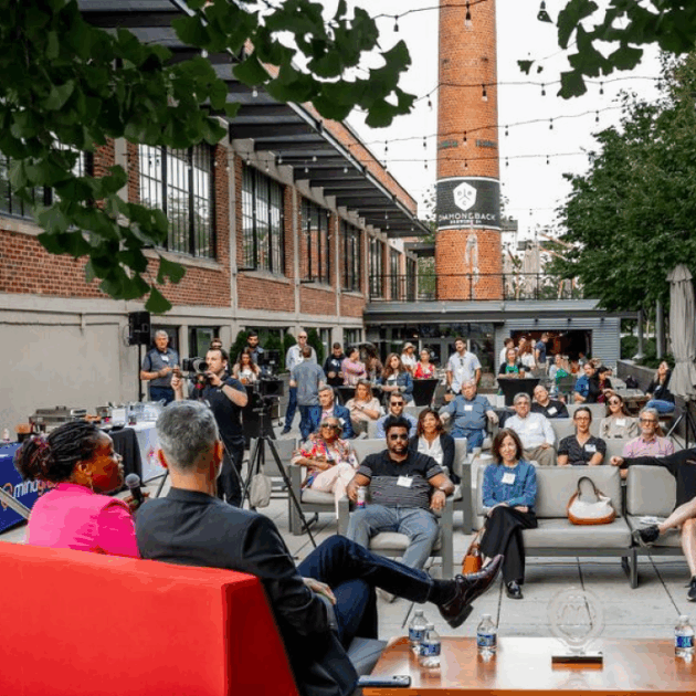 A man and a woman speaking to an audience in an outdoor courtyard.