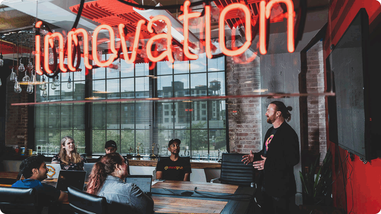 Five people sit at a conference room table and face one person standing at the head of the table talking to the group.