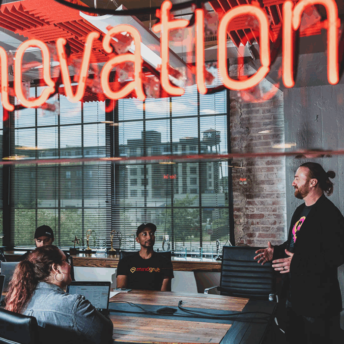 Five people sit at a conference room table and face one person standing at the head of the table talking to the group.