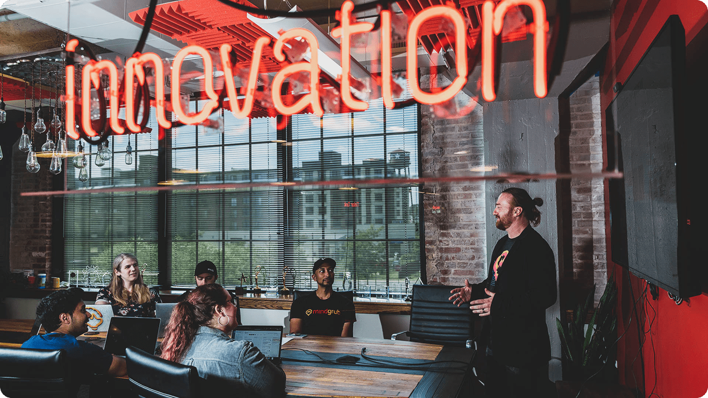 Five people sit at a conference room table and face one person standing at the head of the table talking to the group.