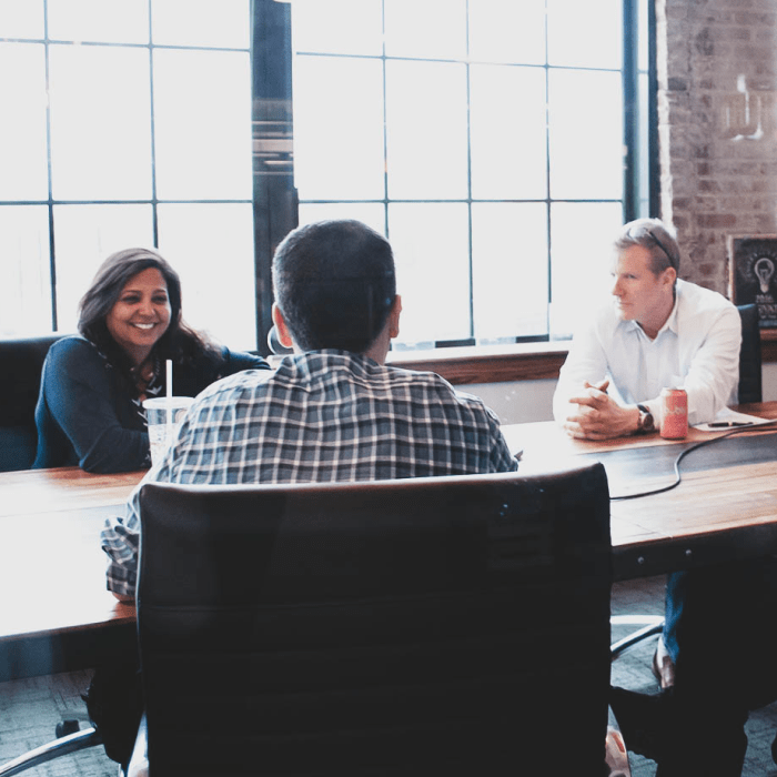 Three people sitting at a conference table. One person's back is to the camera. Two people face the camera smiling.