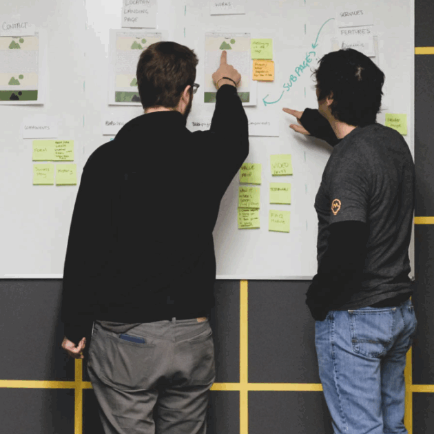 Three people looking at papers and notes on a white board, two of the people are pointing at a specific page on the right