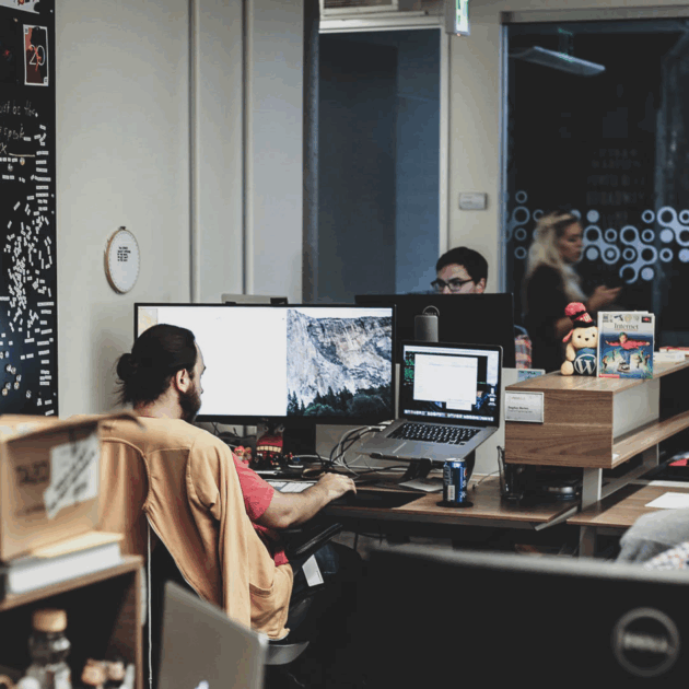 A man on a computer in an office. Another man faces him on a computer.
