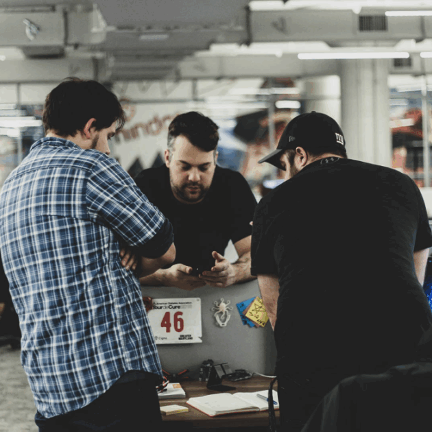 3 men huddled over a phone in discussion. Two men are facing away and one man is facing towards the camera while holding the phone.