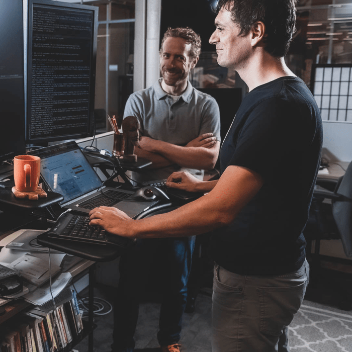 One man stands at his desk, facing his computer and smiling. Another man looks at him from the background and is smiling.